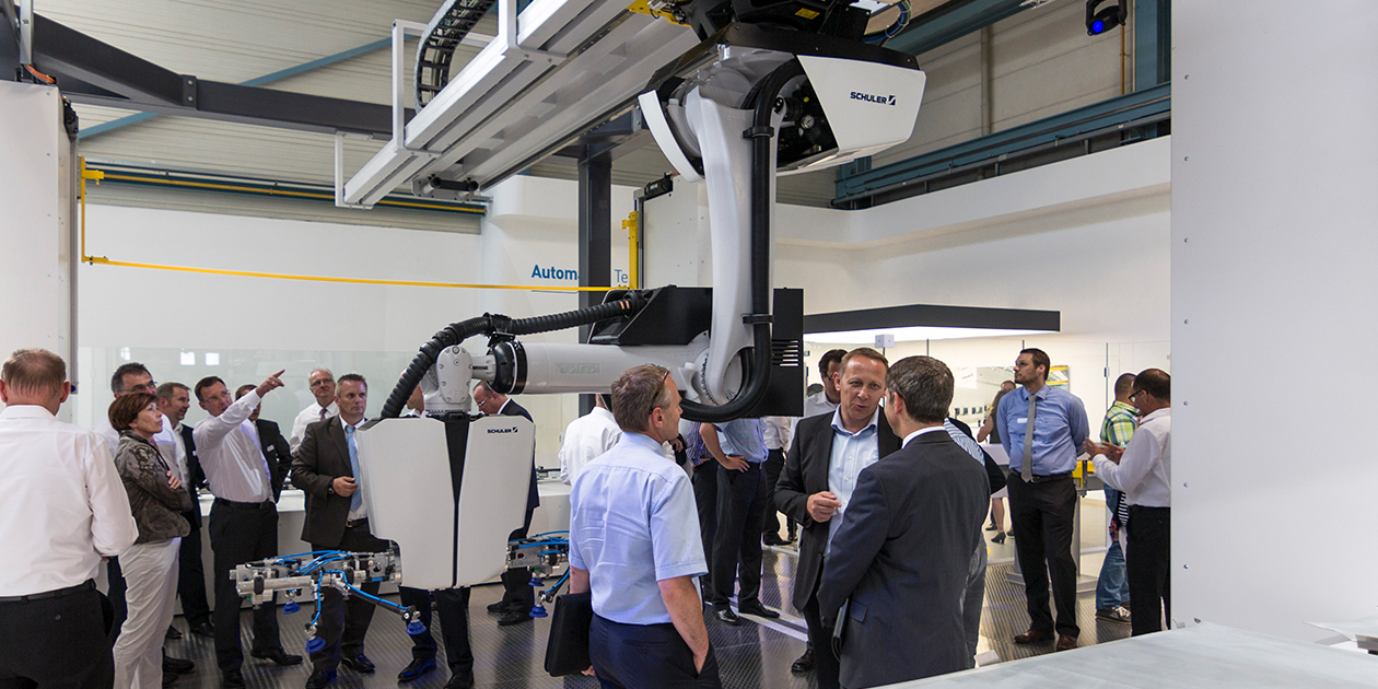 Great interest in the presentation of the Crossbar Robot 4.0 from Schuler in their presentation room. The robot, designed with surfaces in white and black, hangs on a portal guide in the middle of the room. The audience walks around the robot, interested and chatty. Schuler Group - designship GmbH - product design - industrial design - machine design - interface design - iF world design index - Top 25 Industry - Top 100 design studios worldwide - we love design